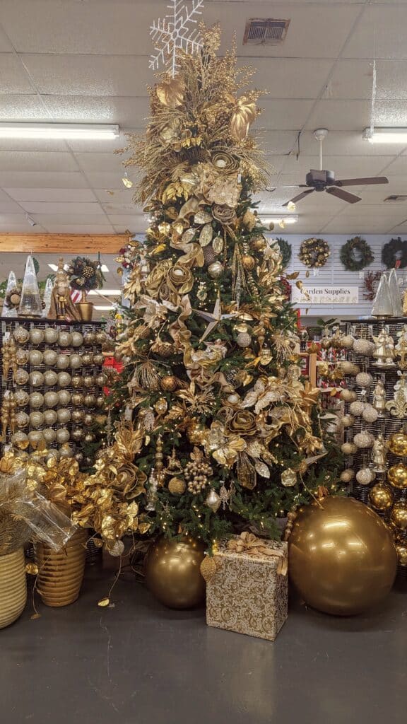 Artificial Christmas Tree with a golden theme. The tree is decorated with golden ornaments, golden ribbons, golden floral picks or stems, among other golden pieces. Surrounding the base of the tree are presents with sparkling golden wrapping paper, golden bows, and oversized golden ornaments, as well as other various golden pieces.