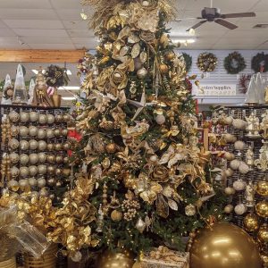 Artificial Christmas Tree with a golden theme. The tree is decorated with golden ornaments, golden ribbons, golden floral picks or stems, among other golden pieces. Surrounding the base of the tree are presents with sparkling golden wrapping paper, golden bows, and oversized golden ornaments, as well as other various golden pieces.